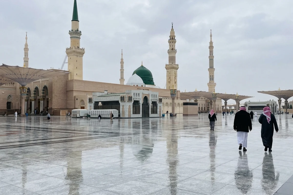 The Green Dome and minarets of Masjid an-Nabawi under an overcast sky, seen across a wet marble courtyard