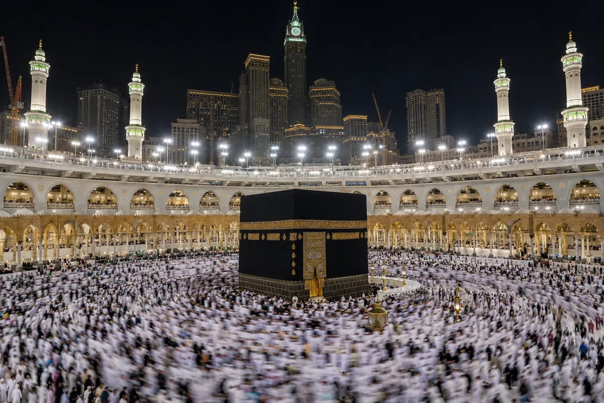 The Kaaba at night surrounded by a dense crowd of pilgrims, with lit minarets behind