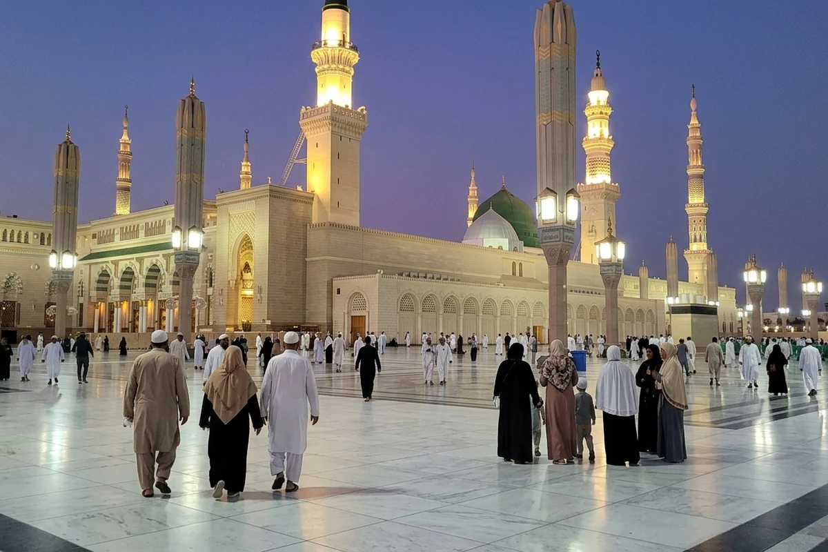 Families walking across a mosque courtyard at dusk beneath lit minarets